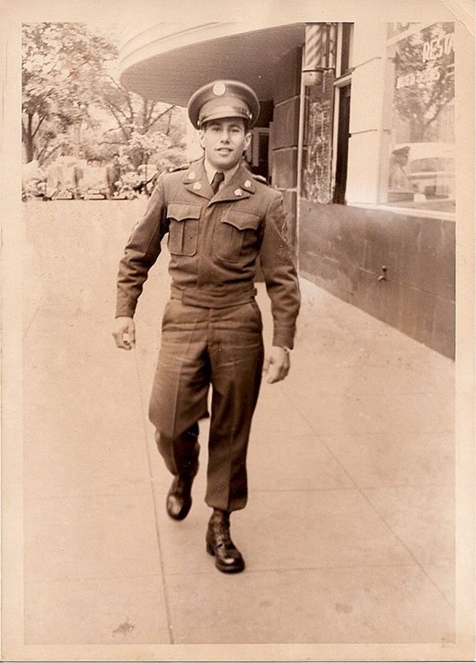 A vintage sepia photograph of a young man in military uniform walking down a city street after the war.